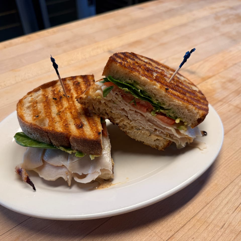 Grilled turkey sandwich with lettuce, tomato, and avocado on toasted bread, cut in half on a white plate.